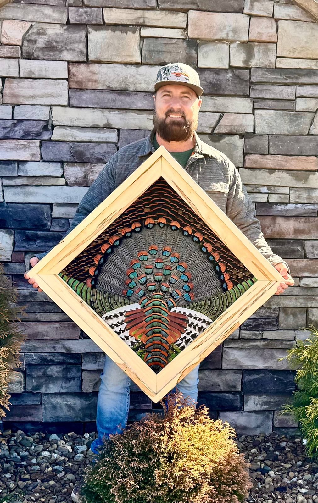 Mark Cooke smiling in front of a stone wall, holding an ocellated turkey fan in a maple diamond frame