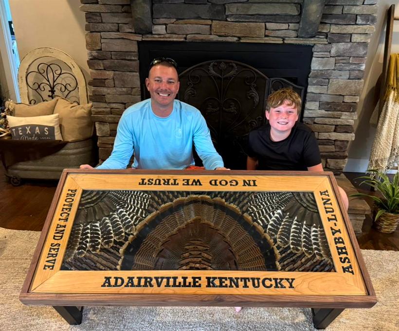 Commissioned coffee table with turkey feathers under glass and an engraved border reading Adairville Kentucky, a father and son seated behind it
